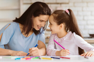 A mother and her daughter are seated at a table, both smiling and drawing with colorful pencils. They appear to be enjoying a fun and creative activity together.