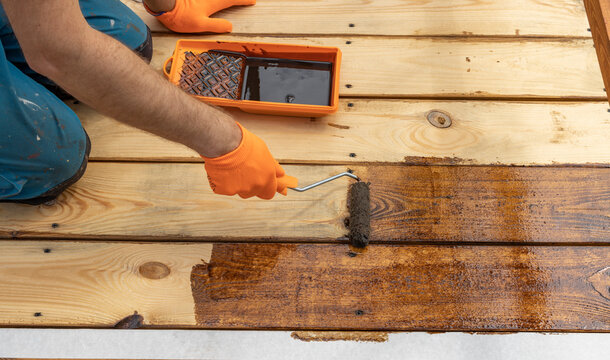 A worker wearing orange gloves carefully applies a dark brown stain to a wooden deck with a roller. The deck is partially stained, with the freshly stained wood gleaming in the sunlight.