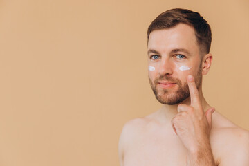 Obraz premium Attractive bearded man with healthy skin, applies cream moisturizer for anti wrinkle or anti aging, cares of body, poses naked or shirtless, isolated over beige background with free space.