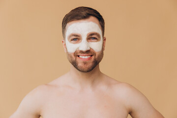 Fototapeta premium Beauty portrait of shirtless unshaved smiling man applies facial care mask against beige studio background. Dermatology health, luxury spa procedures. Concept of male beauty