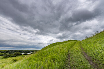 A winding path through a field of tall grass leads to a grassy hilltop under a cloudy sky..
