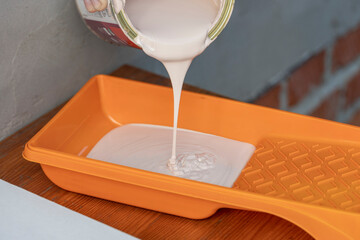 A can of white paint is being poured into an orange paint tray, preparing for a home improvement...