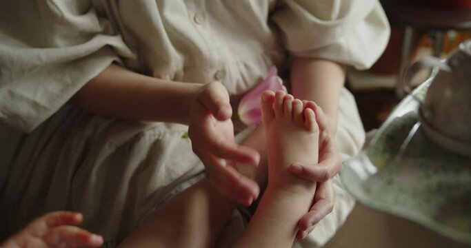 Young loving mother stroking foot of her adorable little daughter sitting near. Close-up slow motion shot.