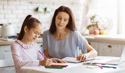 Obraz premium A young girl sits at a kitchen table with her mother, intently coloring with crayons. There is a notebook and laptop on the table, suggesting that they are doing some kind of creative activity