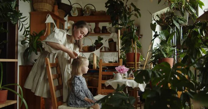 Young loving mother tying a hair bow for her adorable little daughter in grey dress sitting on a wooden chair among decorative plants in the home garden. Wide slow motion shot.