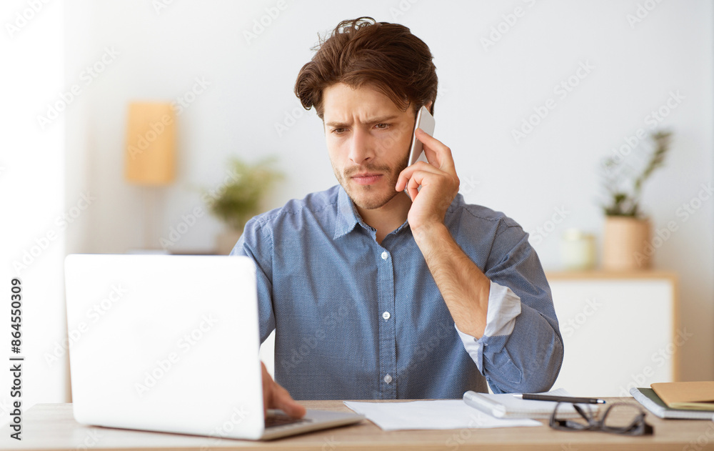 Poster a man sits at a desk talking on his phone while working on his laptop. he looks focused and serious.