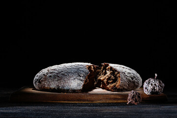 Broken brown rye round bread with crumpled paper dried shriveled fruit in dark on a wooden board. Selective focus
