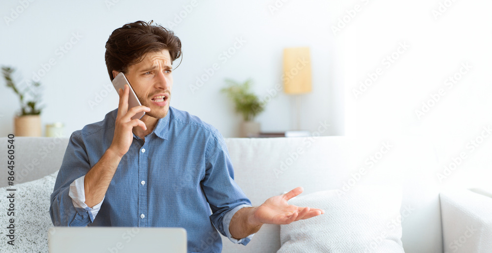 Leinwandbilder a frustrated young man talks on the phone with a stressed look on his face while sitting on a couch.