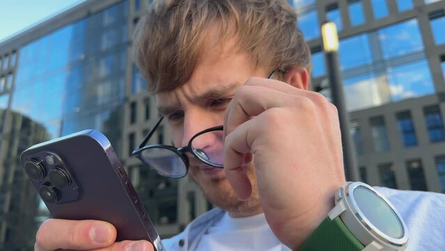 Young man with myopia wearing eyeglasses with eyesight problems trying to read phone text outdoors