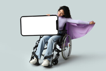Beautiful young happy African-American woman in wheelchair and superhero cape with blank poster on grey background