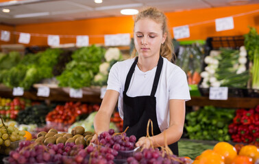 Fifteen-year-old girl who works part-time in a store as a trainee saleswoman puts ripe grapes on the counter, laid out..in boxes