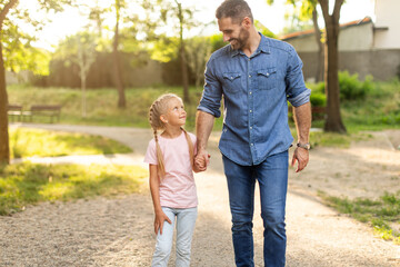 A father and his young daughter walk hand-in-hand on a gravel path through a park. The sun shines...