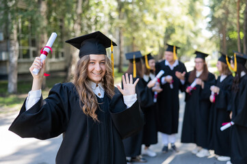 Group of happy students in graduation gowns outdoors. A young girl with a diploma in her hands in the foreground.