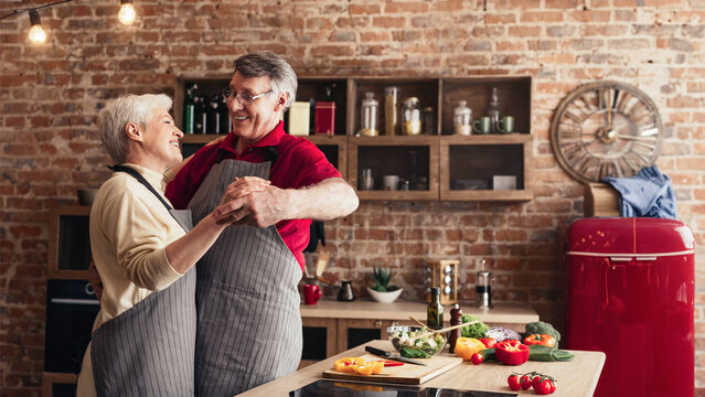 A senior couple in aprons, smiling and holding each other, are dancing in a kitchen. Fresh vegetables are on the counter in front of them. - Powered by Adobe