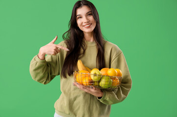 Pretty young woman pointing at fresh fruits in basket on green background