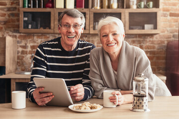 A happy senior couple smiles for the camera while sitting at a kitchen table, enjoying coffee and using a tablet. The man is wearing a striped shirt, and the woman is wearing a grey sweater.