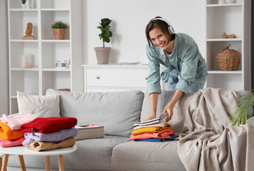 Young woman in headphones folding different clothes at home