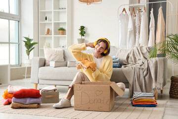 Young woman in headphones packing baby clothes in donation cardboard box on floor at home