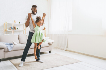 A happy black father lifts his young daughter in the air as they play in their bright living room....
