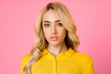 Attractive young woman with long, blonde, wavy hair is wearing a yellow shirt against a pink background. She looks directly at the camera with a neutral expression.
