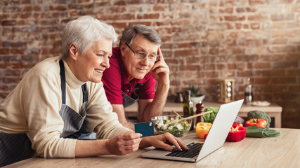 A senior couple is happily using a laptop to order groceries from their kitchen. They are both...