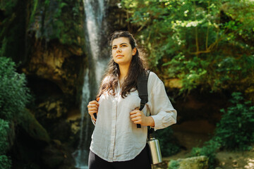 One young caucasian woman is taking a break from hiking near the river and waterfall in the forest	