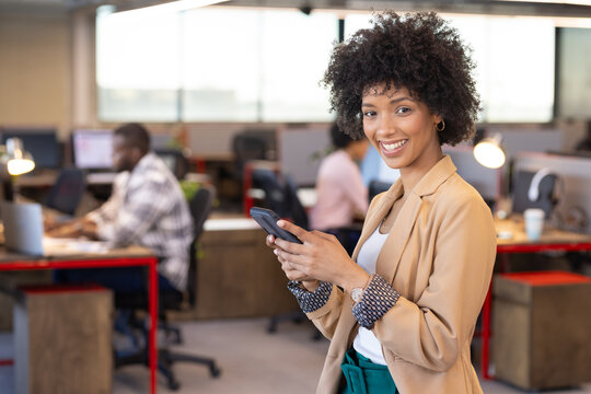 Using smartphone, smiling African American businesswoman in modern open office setting