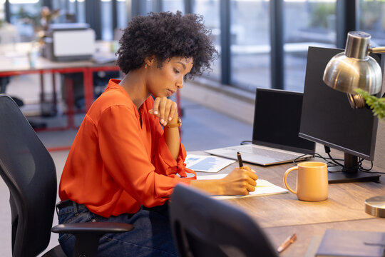 African American businesswoman writing notes at desk with laptop in modern office