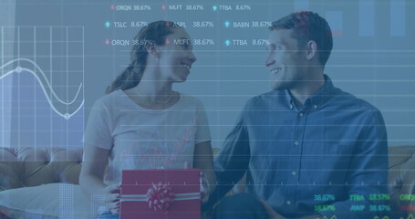 Image of graphs and trading boards, diverse man surprising woman by presenting gift at home
