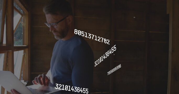 Image of multiple changing numbers over caucasian man standing near window and using laptop