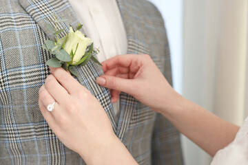 Bride putting boutonniere on her groom's jacket against light background, closeup. Wedding accessory