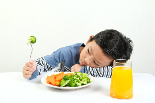 A young boy leans on the table, refusing to eat his vegetables and drink orange juice, displaying boredom and reluctance. Perfect for illustrating picky eating habits in children.