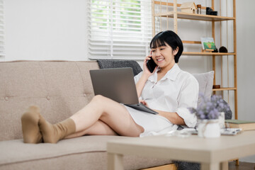 A young woman in casual clothing relaxes on a sofa, multitasking with a phone call and laptop in a...