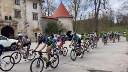A group of cyclists are racing down a street with a crowd of spectators watching. The cyclists are wearing bright red jerseys and helmets