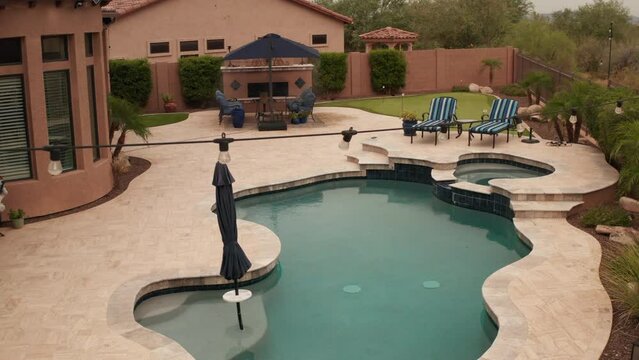 An aerial 4k video of a Deseret landscaped backyard in Arizona featuring a travertine tiled pool deck with a spa, outdoor fireplace, outdoor kitchen and putting green.