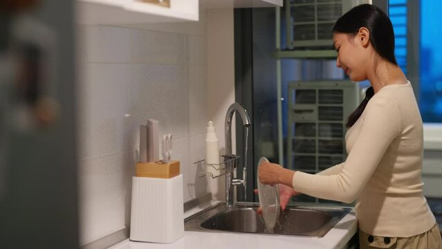 Happy Asian woman do housework washing dishes in the sink after dinner in the kitchen at apartment. Attractive girl using dishwashing liquid and sponge cleaning kitchenware at home kitchen in evening.