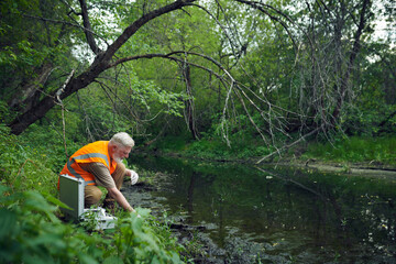 Fototapeta premium Wide shot of senior Caucasian male ecologist in safety vest taking samples at bank of river in forest on summer day, copy space