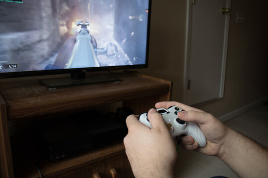 Caucasian male holding white video game controller playing first-person shooter game on TV in bedroom