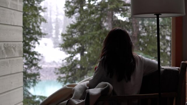Woman Enjoying A Beautiful Snow Day In A Cozy Winter Cabin; Snow Falling Scenery Outside The Window; Winter Forest, Nature, Lake Scenery; 4K.