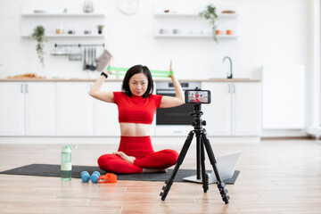 Fototapeta premium Focused woman in red activewear filming fitness tutorial at home using resistance bands. Exercise equipment, water bottle, and laptop nearby, creating realistic workout setting