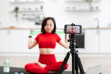 Fototapeta premium Woman in red sportswear filming fitness workout video in home kitchen setting, demonstrating exercises with resistance band, seated in yoga pose, camera on tripod recording session