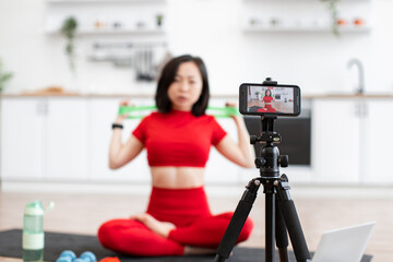 Fototapeta premium Woman in red activewear filming fitness video in kitchen using smartphone and tripod. Demonstrating resistance band exercises during home workout session.