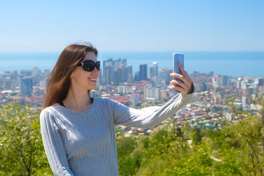 Smiling woman tourist in sunglasses makes selfie on smartphone standing on mountain in Batumi city. Amazing view of Black sea. Travel, tourism, wanderlust, adventure, explore, visiting attractions.