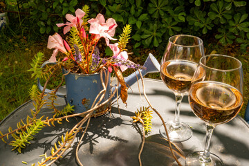 A round garden table in the garden. A decorative flower vase in the shape of a watering can with floral decorations and two wine glasses with rosé on it