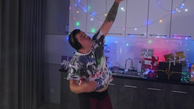 A drugged man with headphones dances waving his arms to music at a home party decorated with colorful garlands.