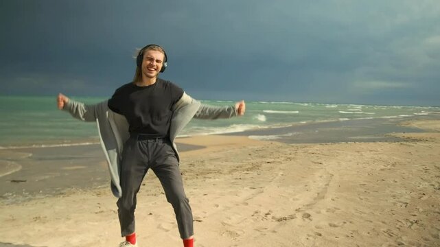 A charismatic guy dances to music on headphones, throwing off his cloak in the wind blowing his hair, cloudy sky and sea waves background.