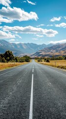 Naklejka premium An open road stretches toward distant mountains under a bright blue sky with scattered clouds, flanked by fields and hills.