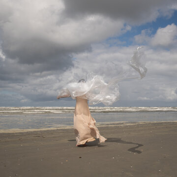 abstract portrait of woman on beach near ocean in long dress holding plastic sheet in the wind resembling a cloud