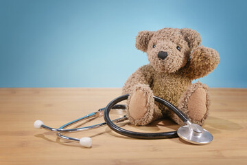 Cute little teddy bear sitting on the table with a stethoscope and waving, to greet the children at the doctor, pediatric medicine and health care concept, blue background, copy space