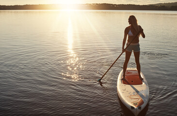 Lake, sunset and woman on paddle board for relax, water sports and vacation on river for tourism. Sunlight, summer and holiday in Germany for female person, swimsuit and travel fitness for adventure